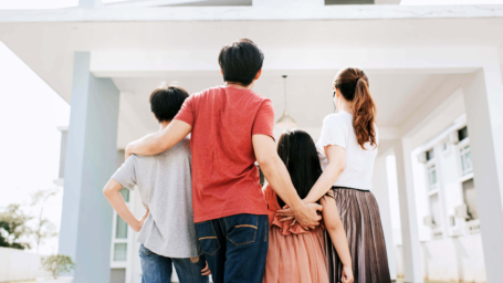 The image shows a family of four standing in front of a modern house, looking up at it. They are seen from behind, with their arms around each other. The family consists of two parents and two children, one older and one younger. The house has a contemporary design with clean lines and a white facade. The image conveys a sense of unity, anticipation, and accomplishment, possibly representing homeownership or a new beginning.
