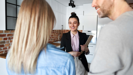 The banner image shows a cheerful real estate agent in a black blazer and pink blouse holding a clipboard while engaging with a couple in a modern kitchen. The couple, seen from behind, appears interested in the conversation. The kitchen features white cabinets and exposed brick, creating a stylish and welcoming atmosphere.